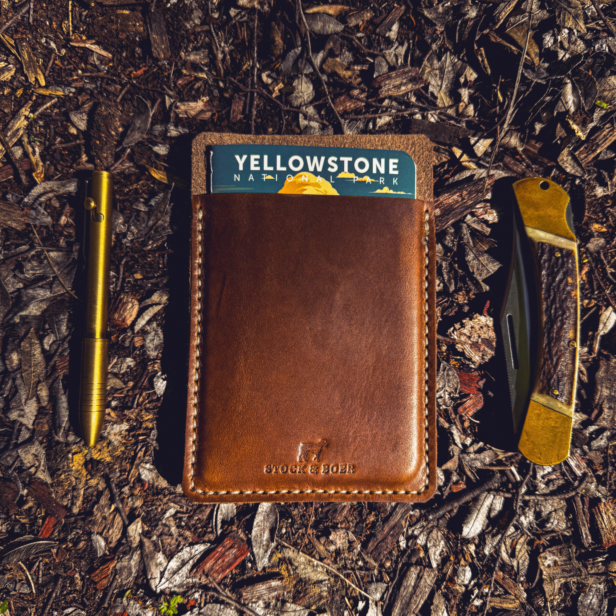 Flat lay of a No. 9 Ledger leather EDC pouch alongside a brass pen and folding knife on a bark and leaf-covered forest floor.