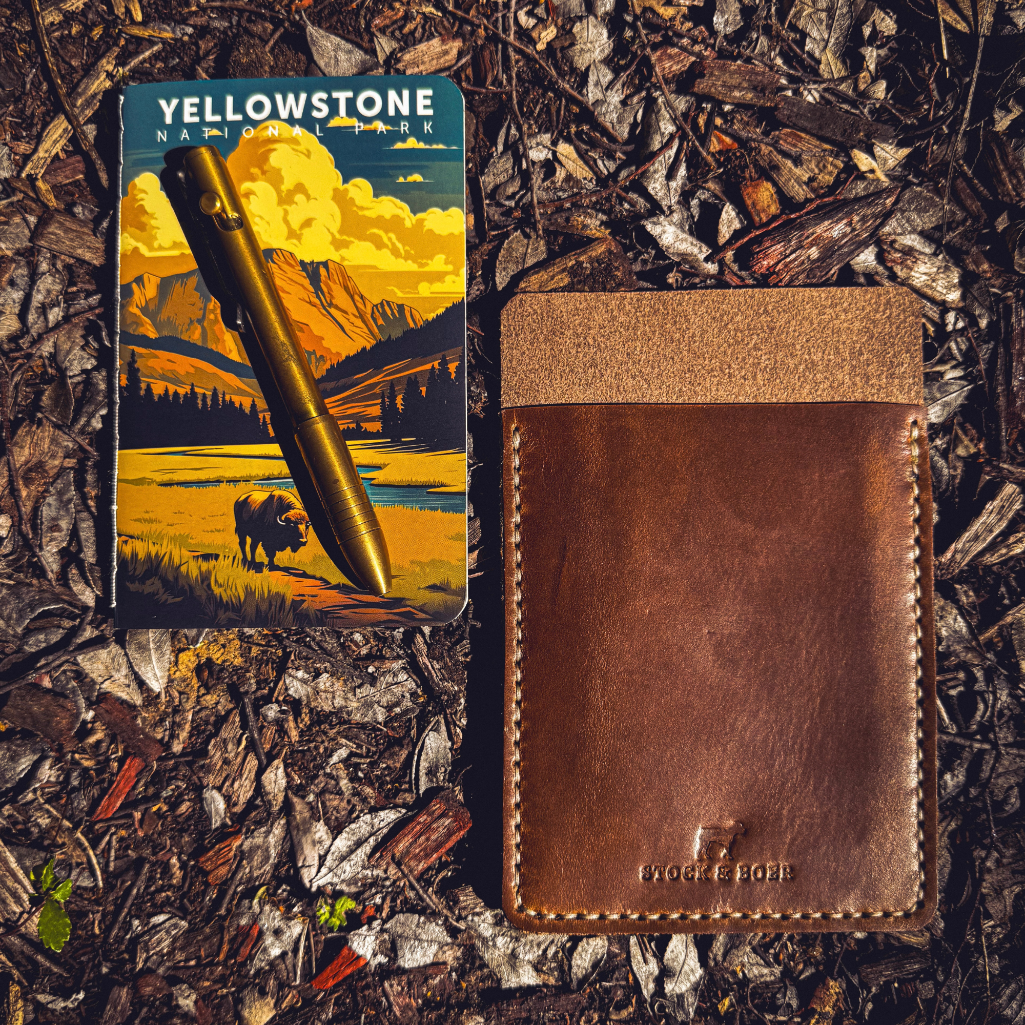 No. 9 Ledger leather EDC pouch positioned next to a brass pen and a Yellowstone National Park illustrated card on a forest ground.