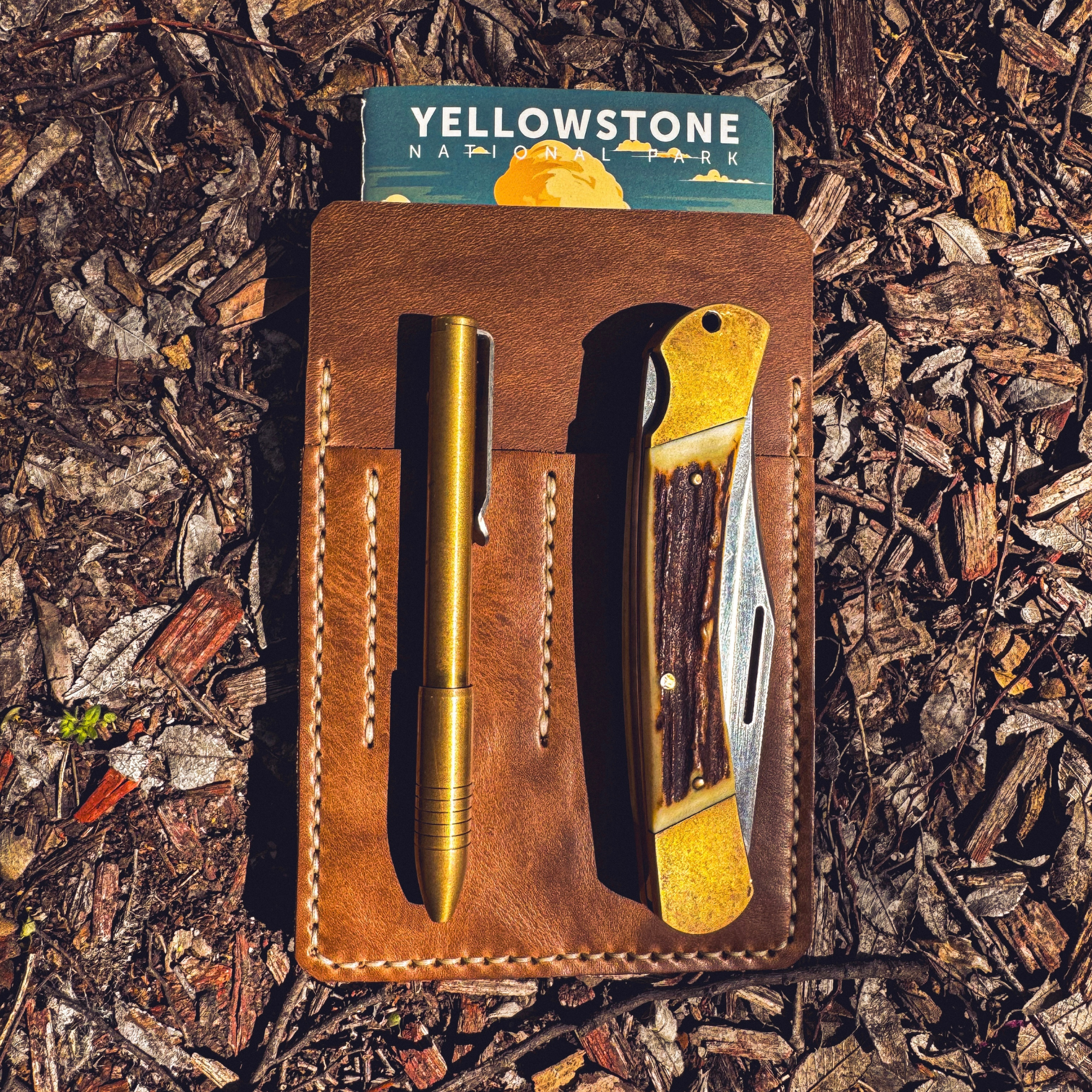 Handcrafted No. 9 Ledger leather EDC pouch in Horween Chromexcel holding a brass pen and pocket knife, displayed on a textured ground backdrop with a Yellowstone National Park notebook.