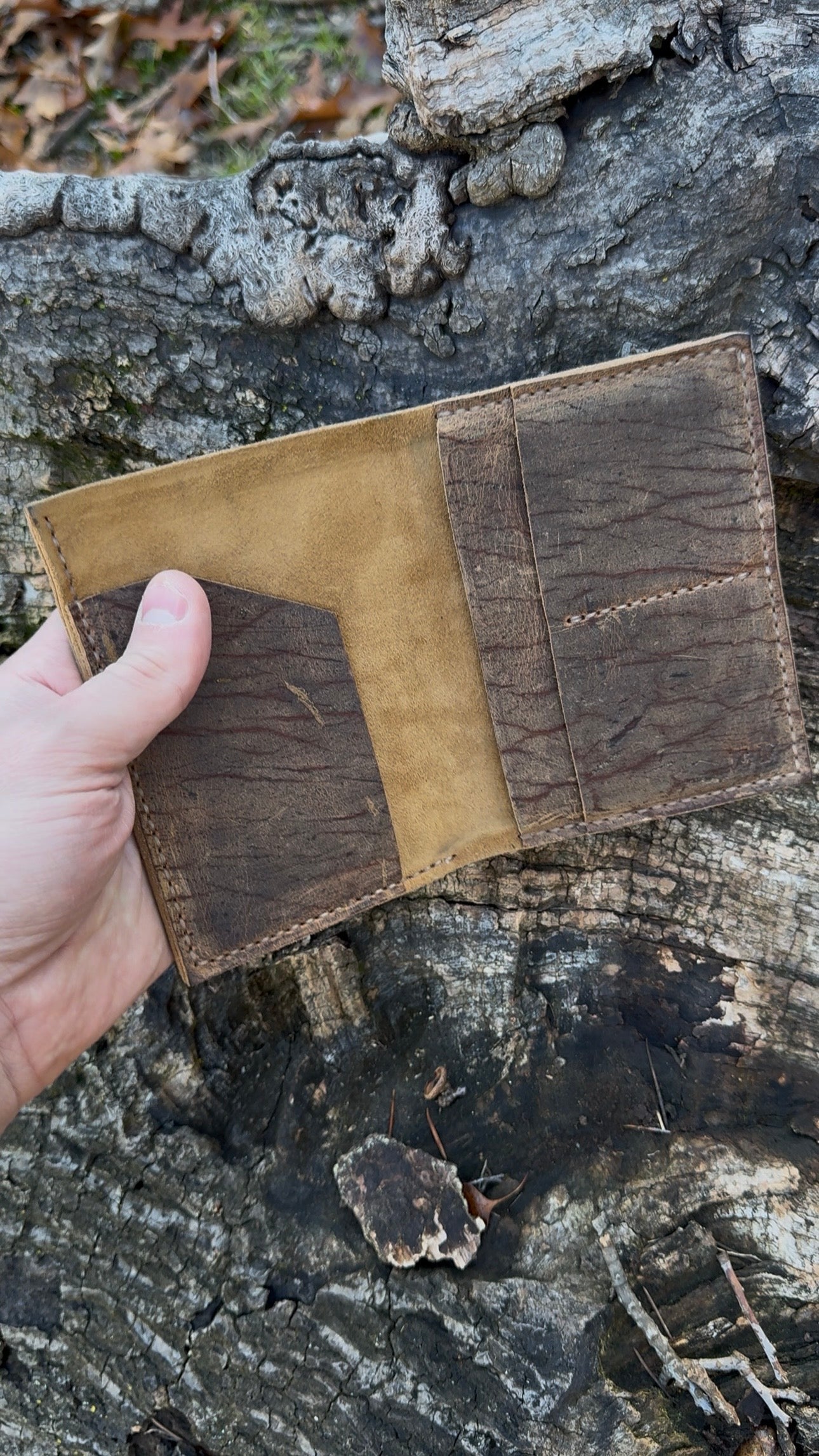 A video showcasing the No. 4 Journeyman Passport Wallet crafted from Baobab Wild African Kudu leather against a rugged tree bark background. The video demonstrates the wallet being turned to show the heavily scarred exterior, opened to reveal the soft golden suede interior lining, and a passport being inserted into the pocket, highlighting the hand-stitched details and texture.