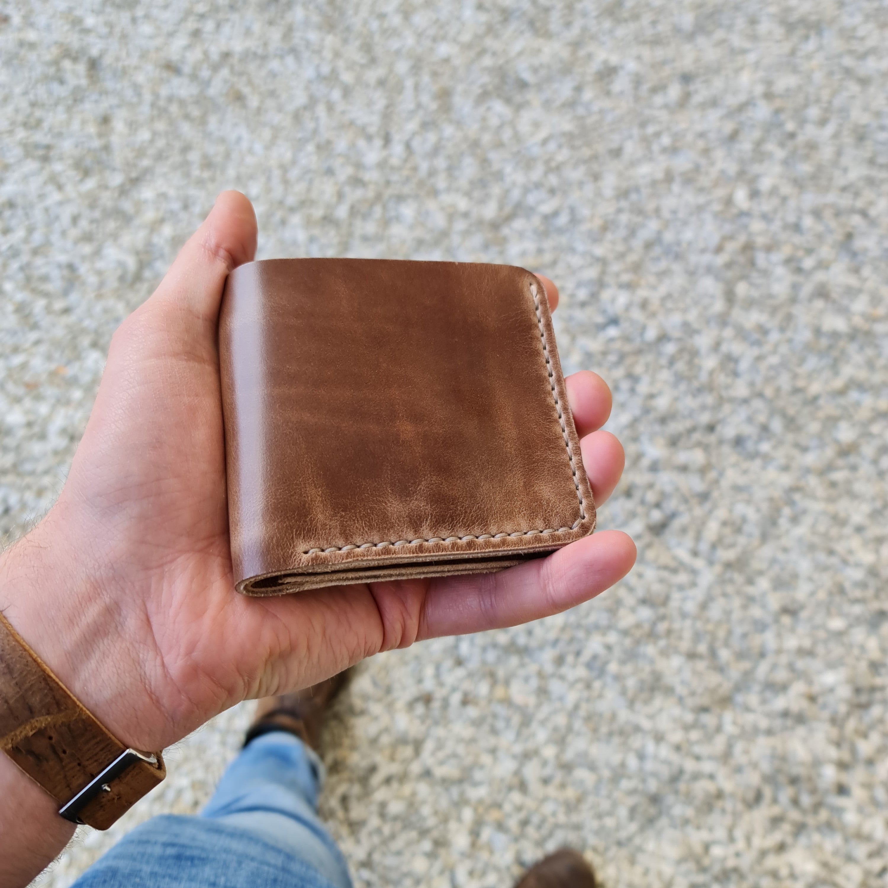 A hand holding the closed exterior of the No. 3 PIONEER Billfold Coin Wallet, crafted from smooth, natural-colored Horween Chromexcel leather. The wallet features neat, light-colored hand-stitching along the edges and is held against a background of blurred gravel and denim jeans.