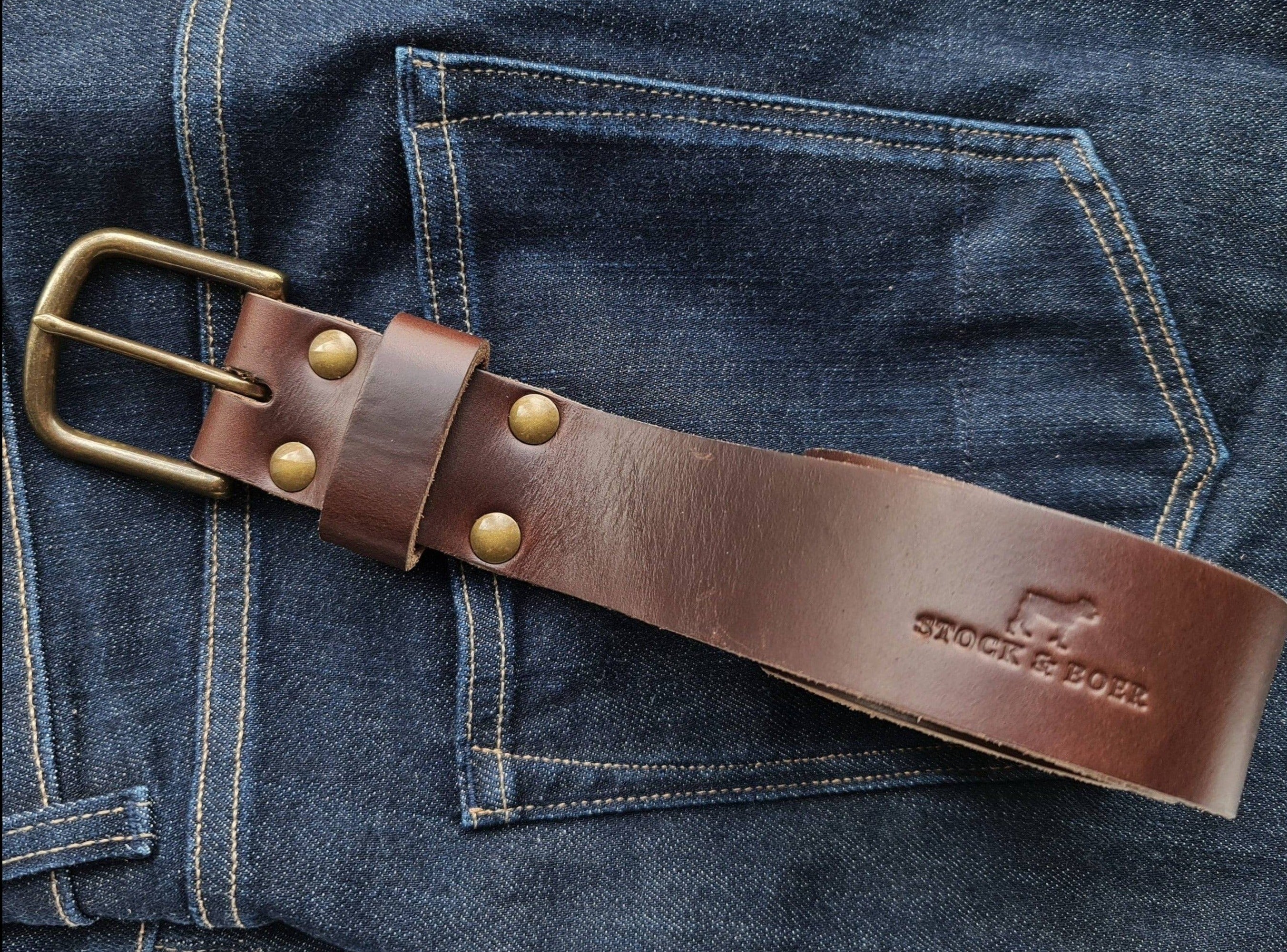 The No. 8 VAQUERO Full Grain Leather Belt in brown Horween Chromexcel leather, displayed against a background of dark blue denim jeans. The belt features a sturdy buckle, brass rivets, and the stamped "STOCK & BOER" logo.