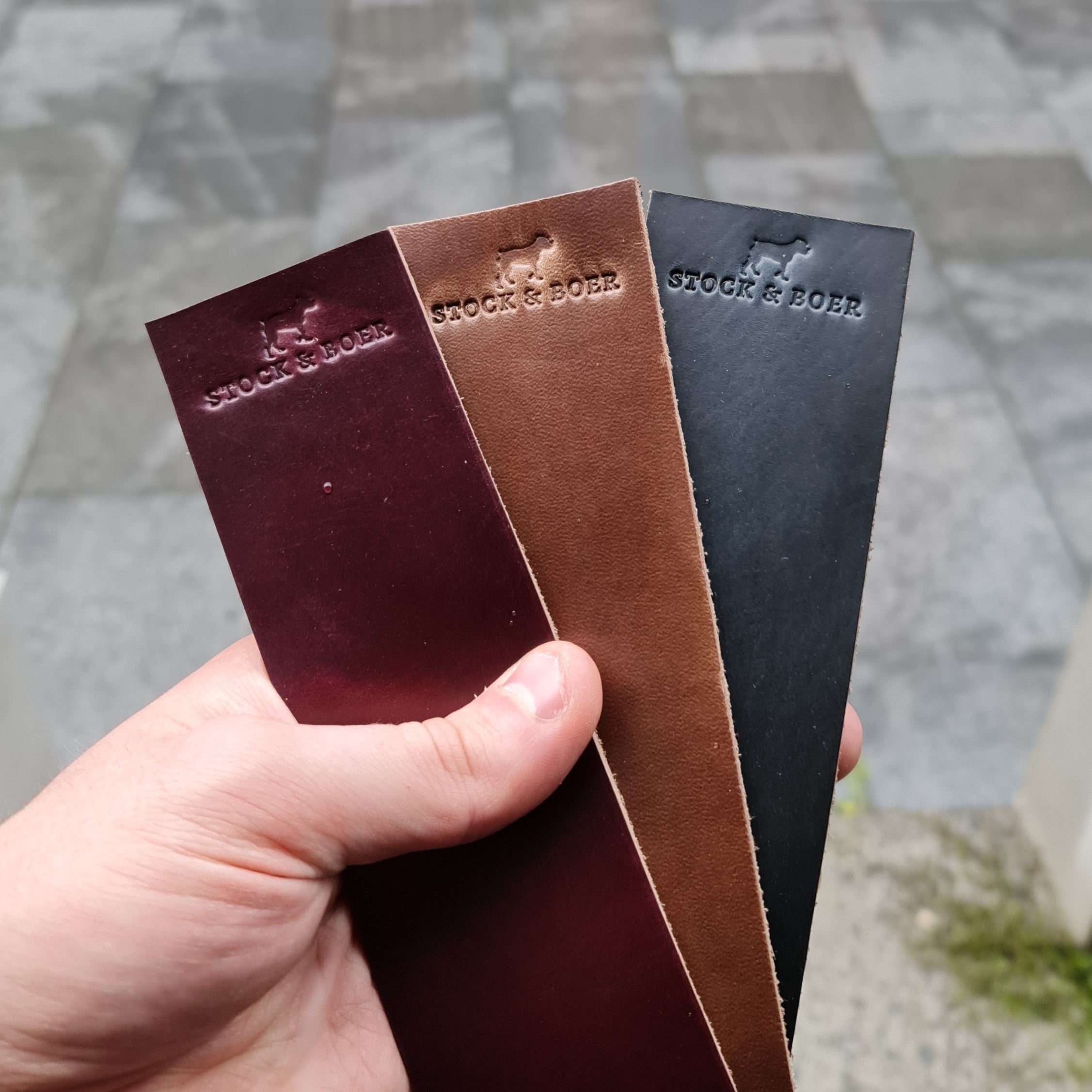 A hand holding three vertical No. 11 RAILMAN Bookmarks crafted from Horween Chromexcel leather, each stamped with the "STOCK & BOER" logo. The bookmarks are displayed in three distinct colors: burgundy, natural, and navy. They are held against an outdoor background of a grey paved stone path.