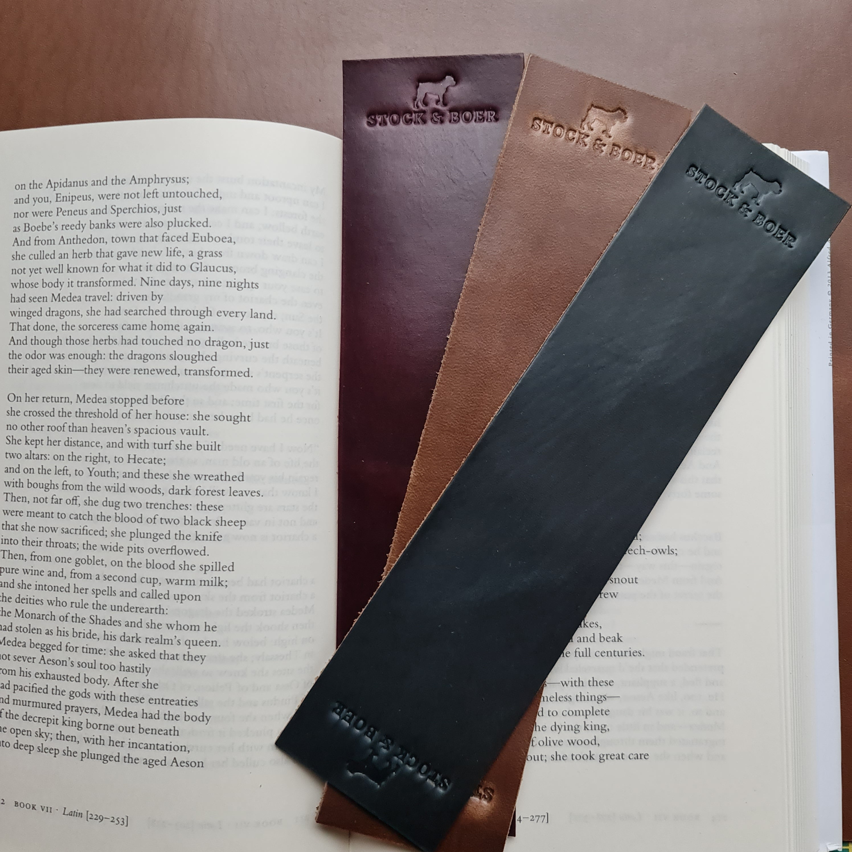 Three No. 11 RAILMAN Bookmarks made from Horween Chromexcel leather rest diagonally across an open book. Each bookmark is stamped with the "STOCK & BOER" logo and features a distinct color: burgundy, natural, and navy. The bookmarks partially obscure the printed text on the open pages of the book.
