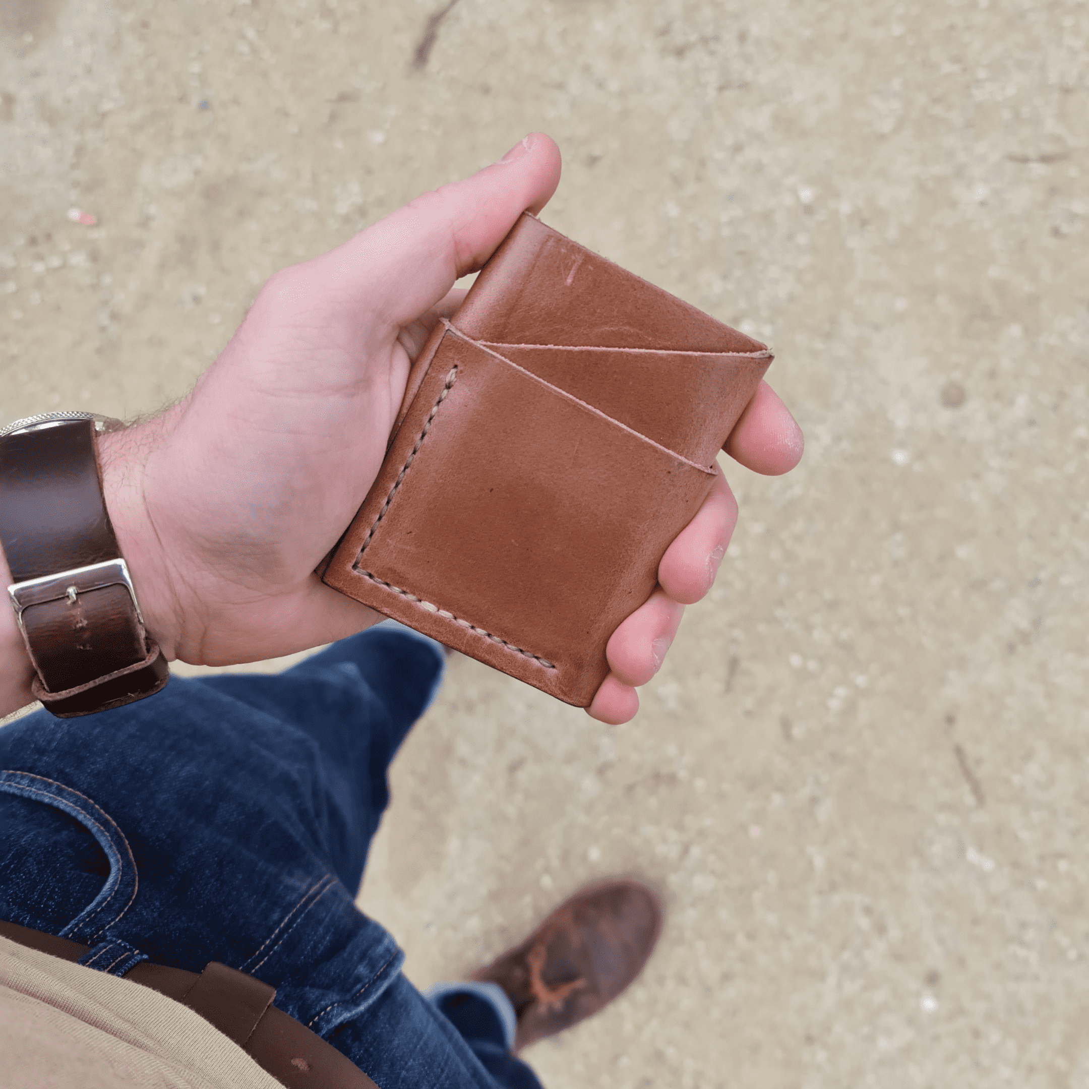 A hand holding the No. 1835 TREKKER Front Pocket Wallet in natural Horween Chromexcel leather. The shot is taken from a top-down perspective, showing the wallet, blue jeans, and brown leather shoes, highlighting the wallet's compact design in an everyday carry setting.