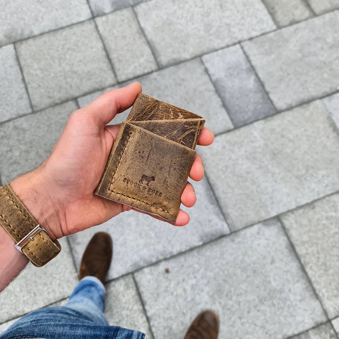 A hand holding the No. 1835 TREKKER Front Pocket Wallet, crafted from rugged, deeply grained Wild African Kudu leather. The wallet features a distinct angled pocket design and is stamped with the "STOCK & BOER" logo. The background shows a grey paved stone surface, with the person wearing matching leather accessories and casual attire.