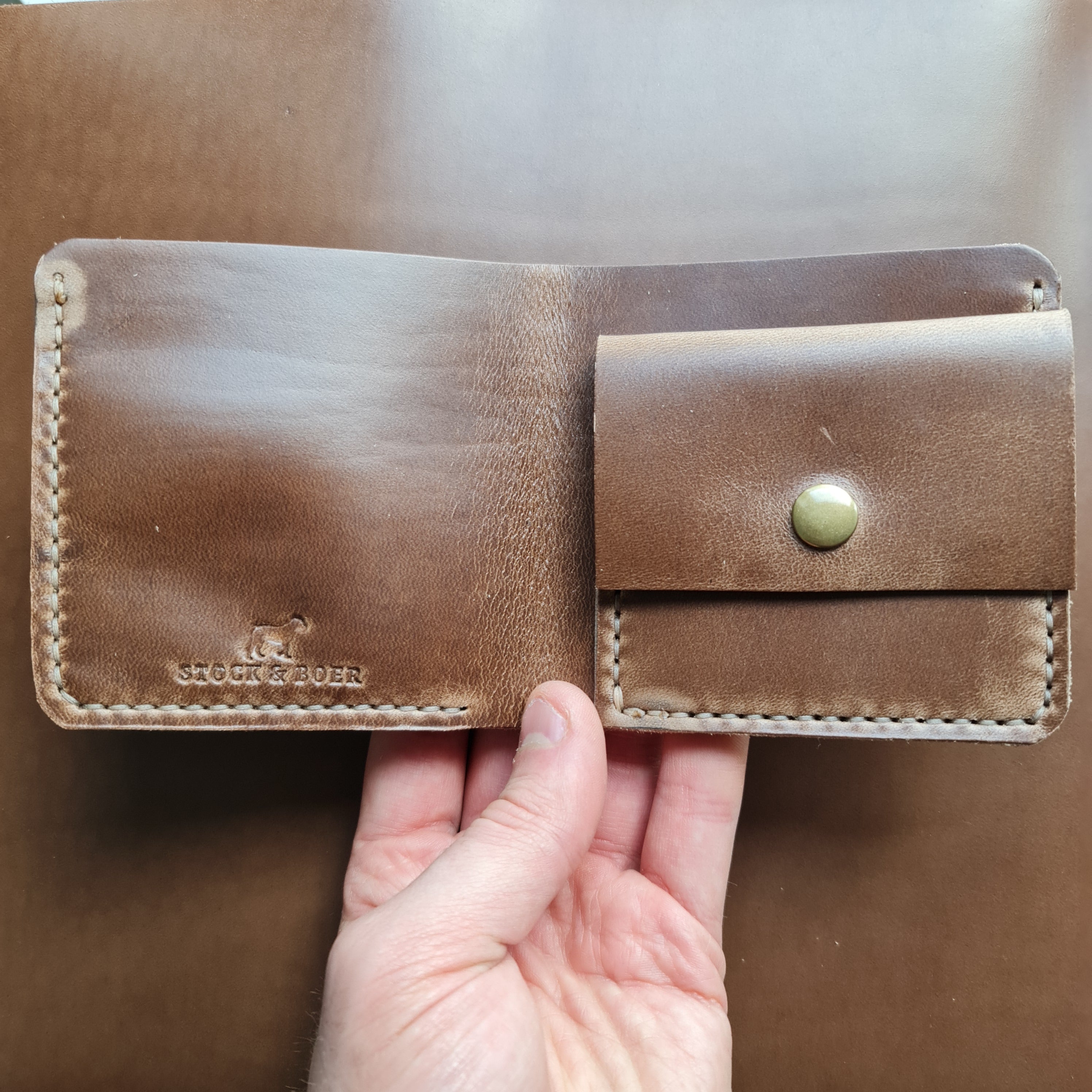 A close-up shot of the interior of the No. 3 PIONEER Billfold Coin Wallet, crafted from natural-colored Horween Chromexcel leather. The open wallet displays a coin pocket secured with a brass snap button and the "STOCK & BOER" logo stamped into the leather. A hand is visible holding the wallet against a neutral, smooth leather background.