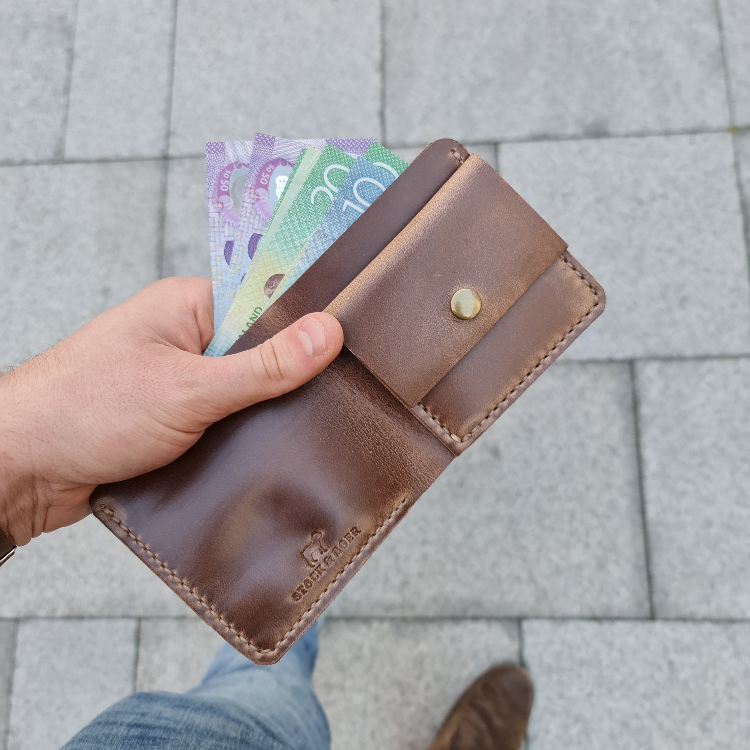 A hand holding the open No. 3 PIONEER Billfold Coin Wallet, crafted from natural-colored Horween Chromexcel leather. The wallet is filled with several Australian banknotes and features a prominent coin pocket secured with a brass snap button, along with a "STOCK & BOER" logo stamped on the front. The wallet is held over a paved stone background with the person's jeans and shoe visible below.