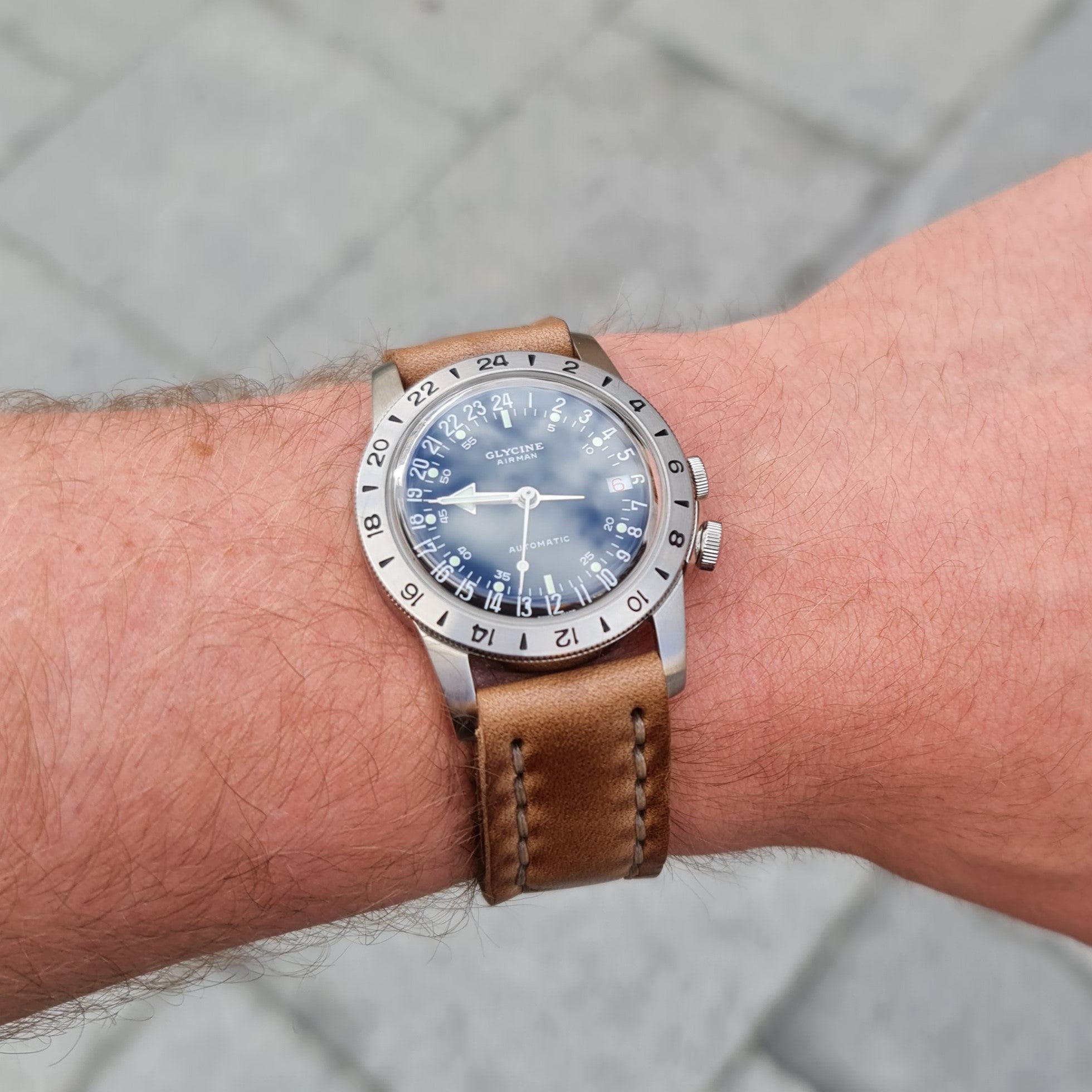 A close-up lifestyle shot of a wristwatch featuring the No. 44 AVIATOR Leather Hand-stitched Watch Strap in natural Horween Chromexcel, worn on a person's wrist. The strap showcases its prominent hand-stitching and the smooth, rich texture of the leather against the backdrop of a paved outdoor surface.