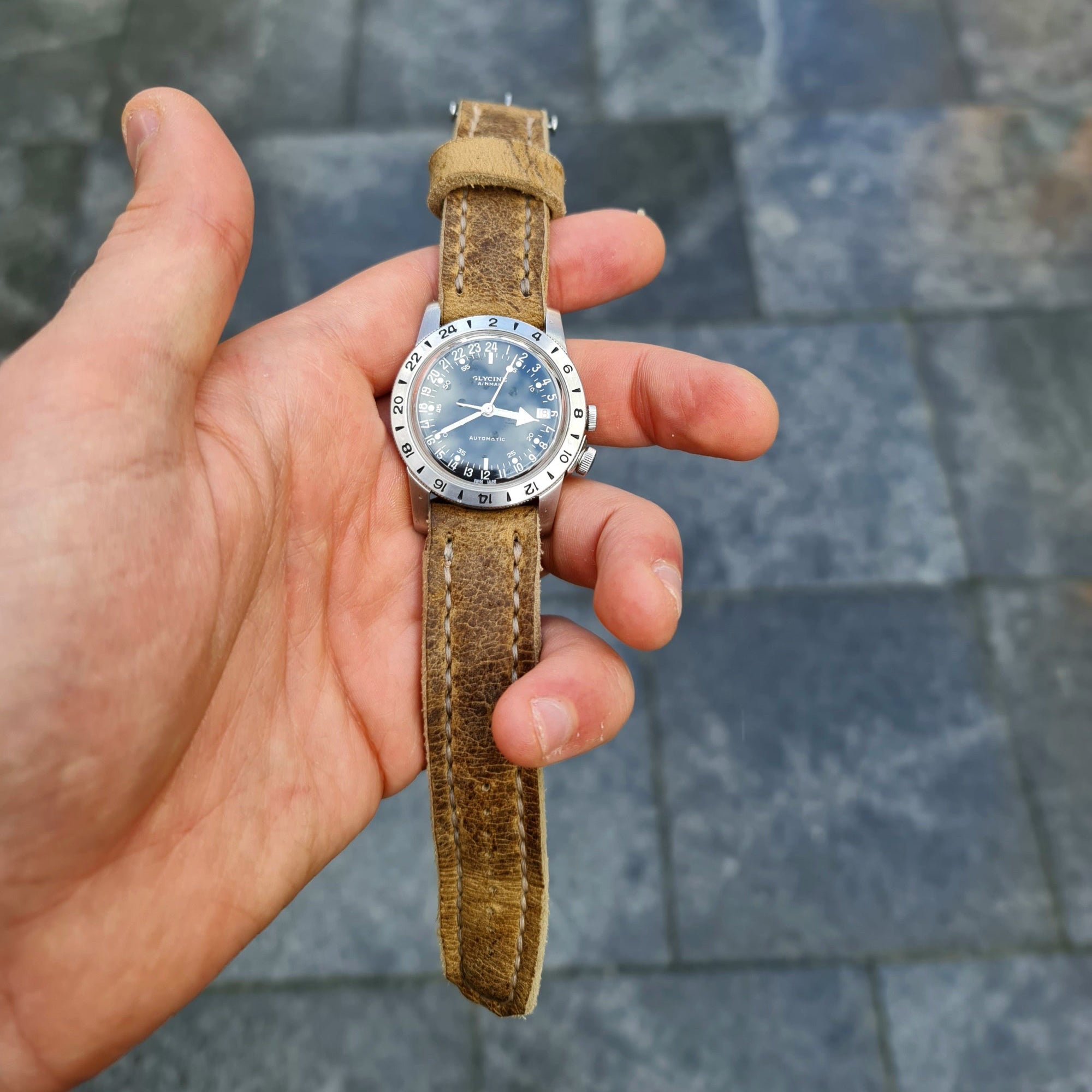 A hand holding a wristwatch fitted with the No. 44 AVIATOR Leather Watch Strap in Wild African Kudu, against a paved stone background.