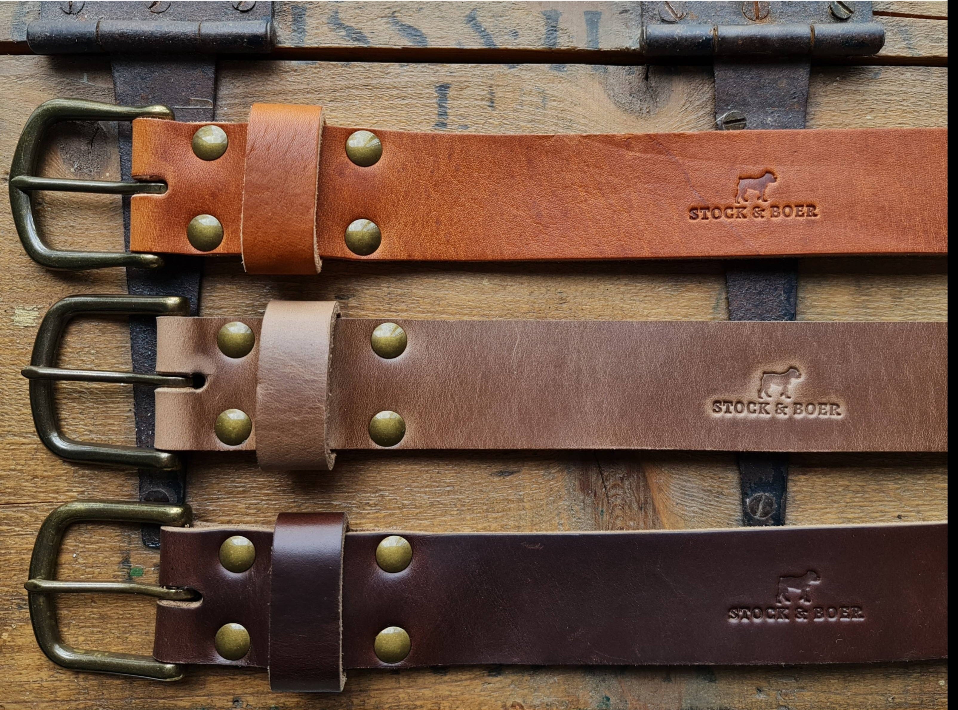 An overview of three No. 8 VAQUERO Full Grain Leather Belts stacked vertically, showcasing the Stock & Boer logo stamp. From top to bottom: Horween English Tan Dublin, natural Horween Chromexcel, and brown Horween Chromexcel, all featuring matching buckles and brass rivets on a vintage wooden background.