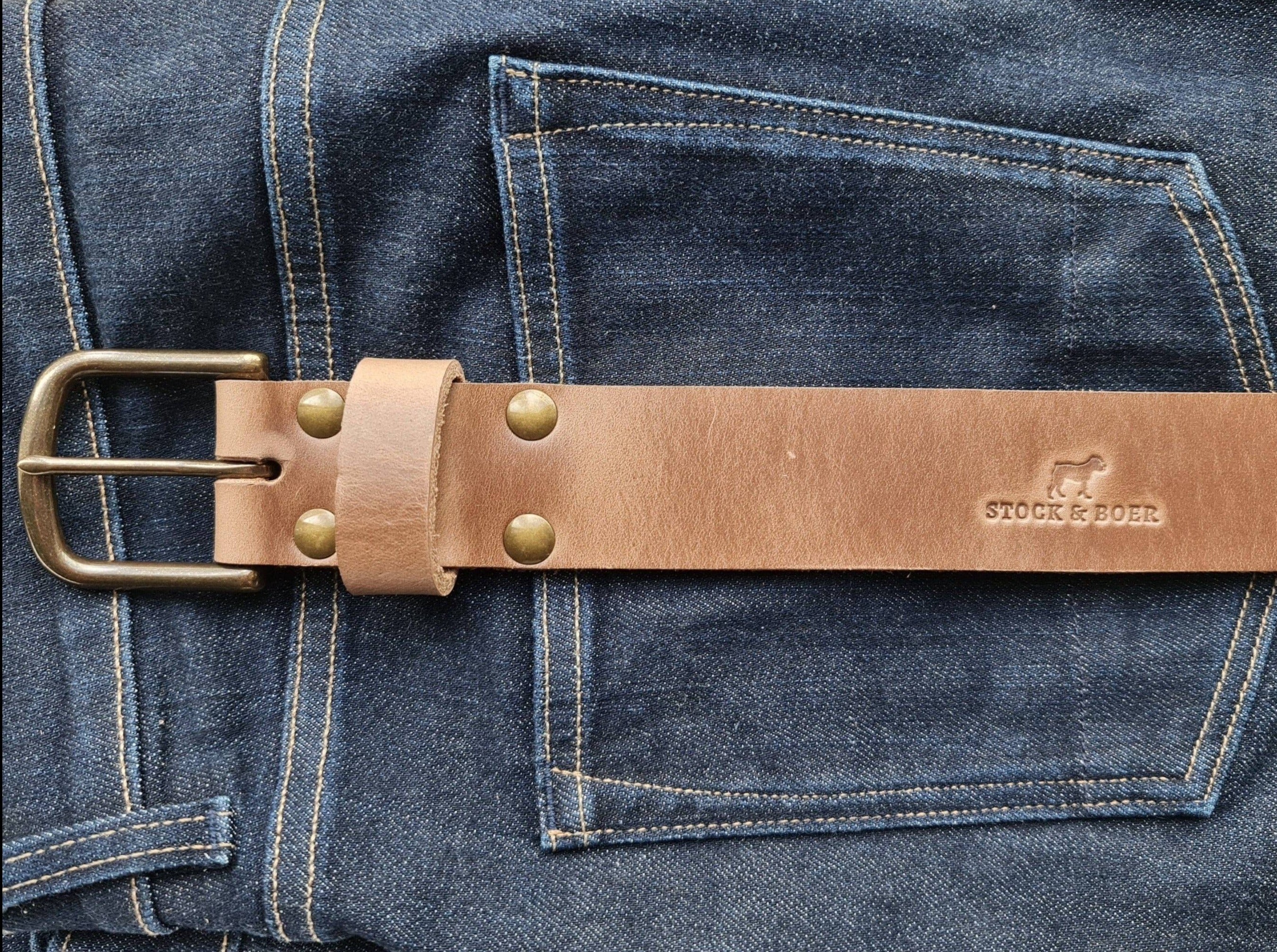 The No. 8 VAQUERO Full Grain Leather Belt in natural Horween Chromexcel leather, displayed against a background of dark blue denim jeans. The belt features a sturdy buckle, brass rivets, and the stamped "STOCK & BOER" logo.